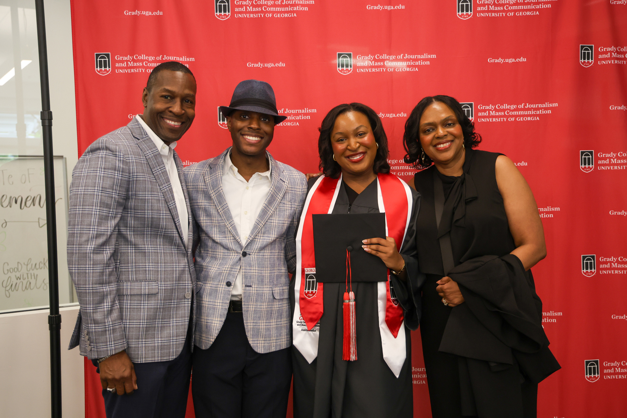 A female student in her graduation gown and holding her cap smiles and poses for a picture with family members in front of a red Grady College step and repeat.
