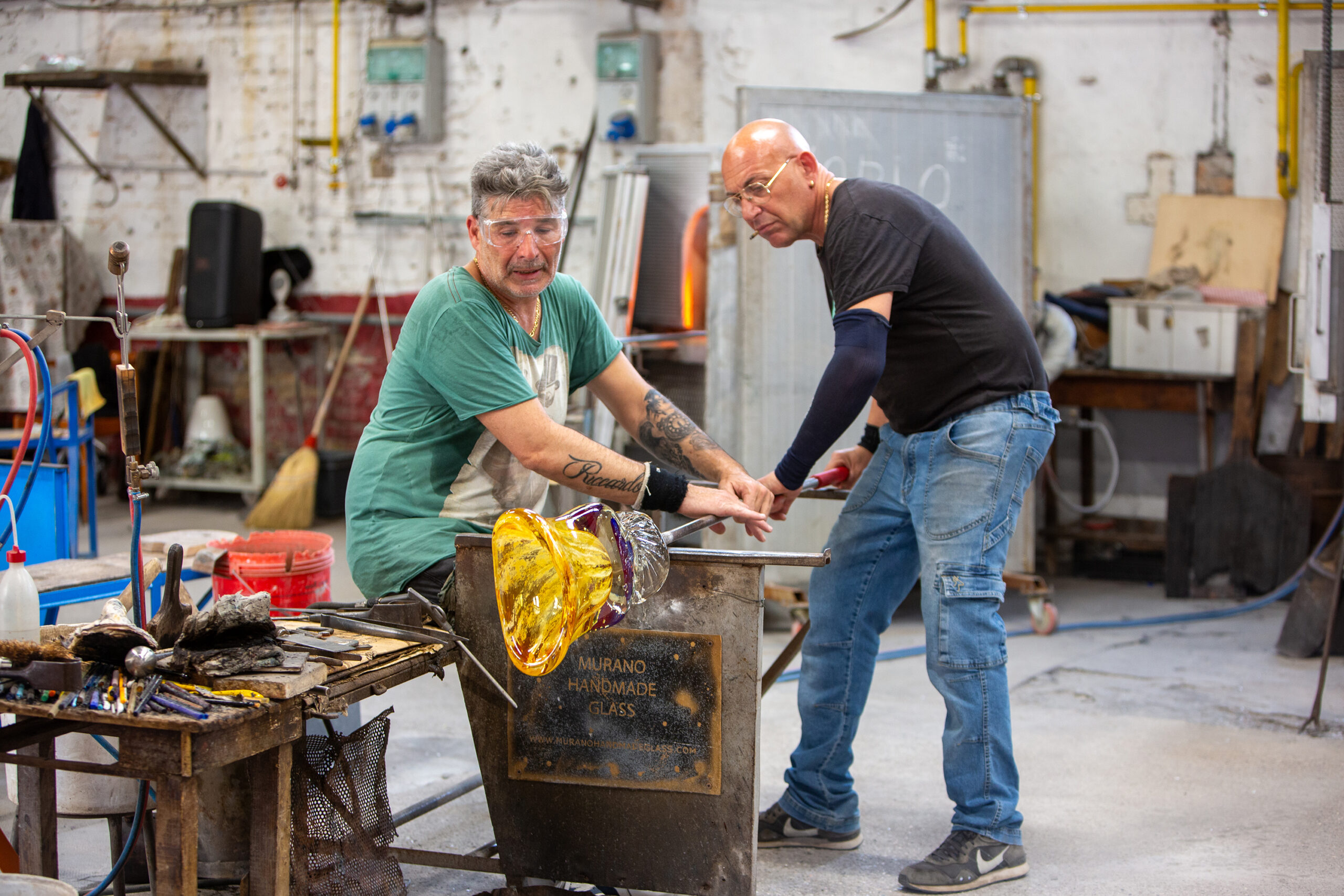 Two men in an workshop hold their hands on a pole over some iron stands and twist a glass at the end of the pole.