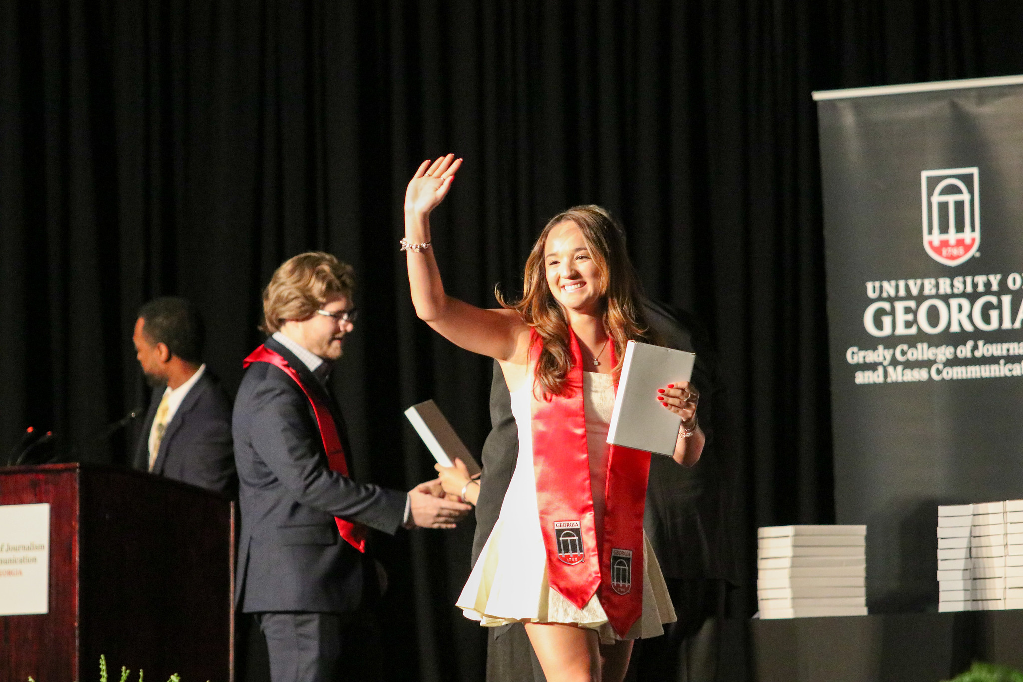 A profile of a female student crossing the stage, smiling and waving. She is wearing convocation stoles and holding a small white box.