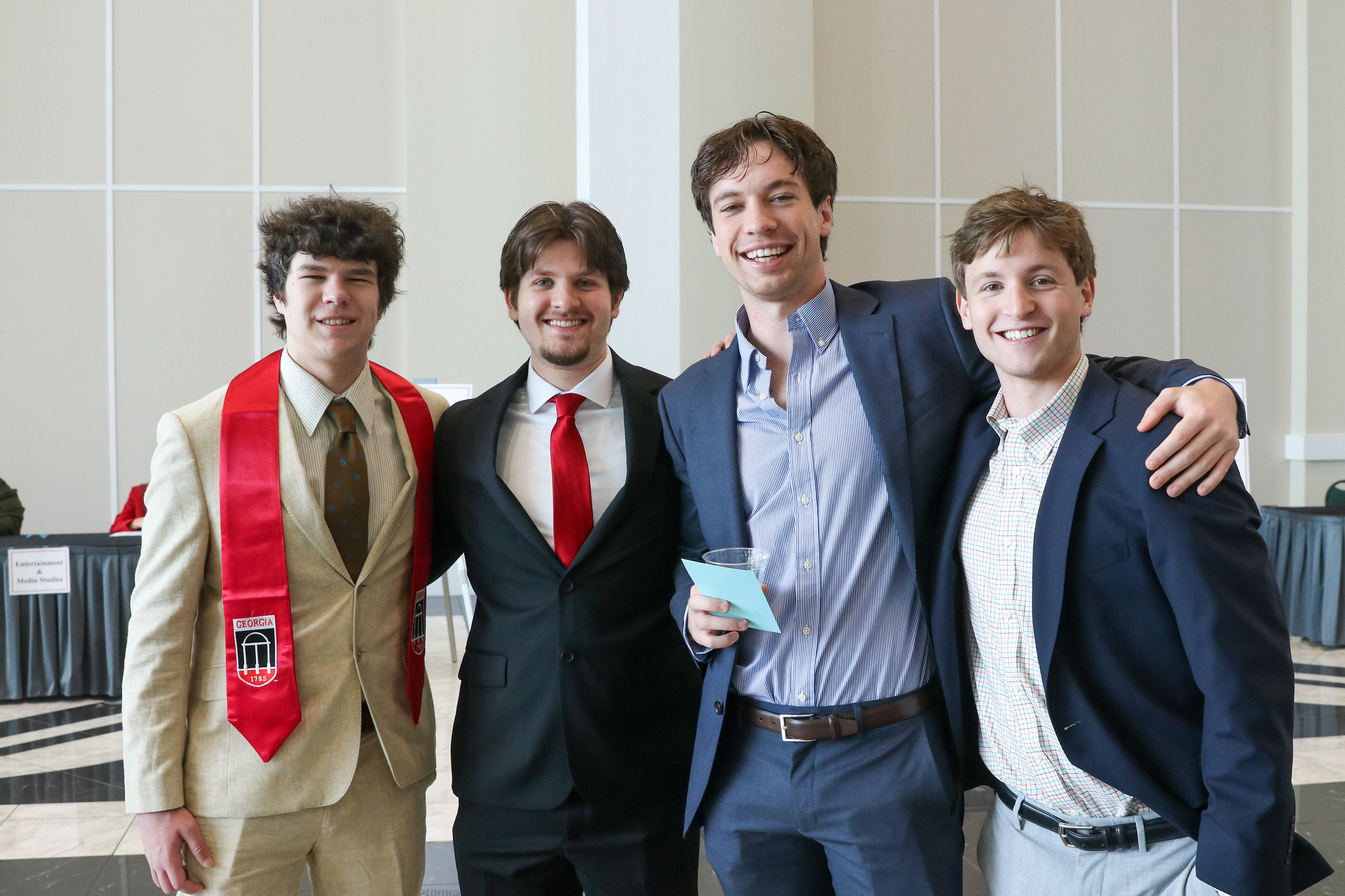 Four male students, all in blazers, smile and pose for a picture. One has a red stole.