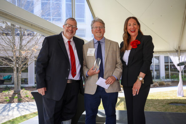 Two men and a woman stand together under a tent. The man in the middle holds a trophy.