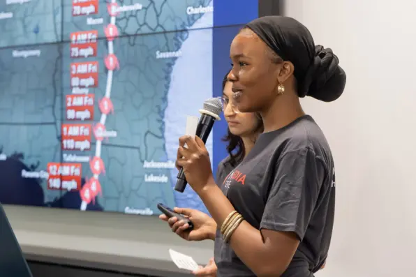 Two students speak in front of a screen depicting a weather forecast on the east coast.