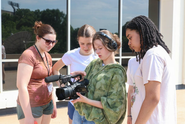 Four young women stand outside, closely looking at a video camera one holds. One wears headphones and a green poncho, and the others watch attentively. There are windows and trees in the background.