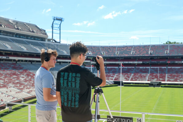 Two young men film with a camera on a tripod in an empty football stadium. One wears headphones, the other adjusts the camera, with the green field and empty red bleachers visible in the background.