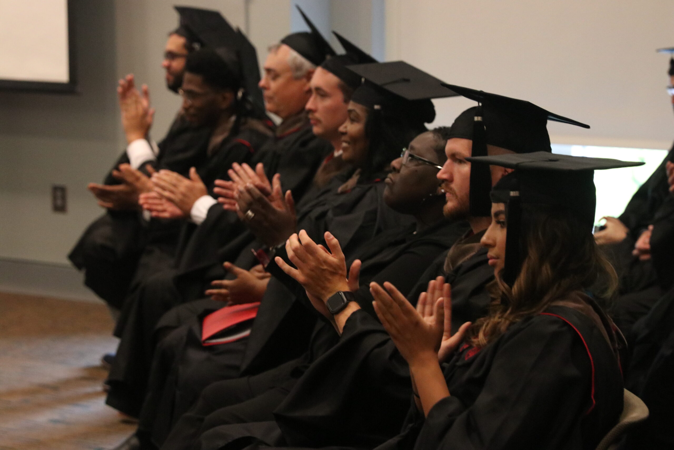 A group of people in graduation regalia sit in a row of chairs applauding.