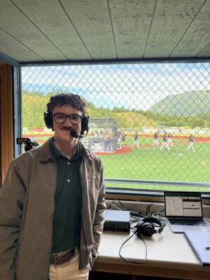Ren Grimsley wears a headset and stands in front of a baseball field.