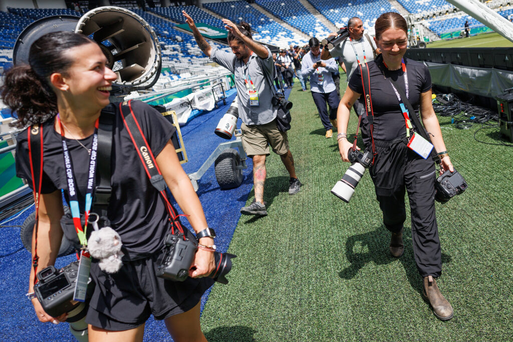 Two female photographers walk in a stadium. They are each carrying two cameras on their shoulders. Several other photographers can be seen behind them.