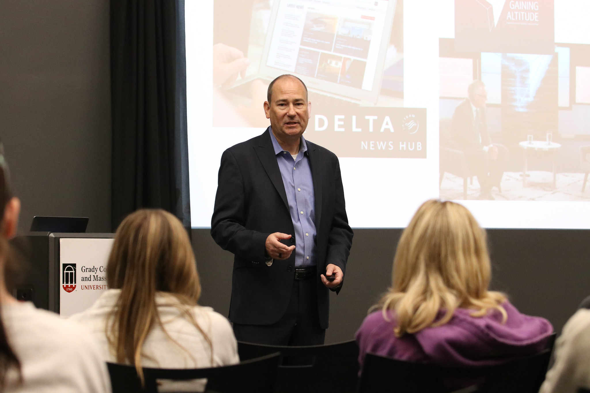 A man gives a presentation to students. A digital projection in the background says Delta.