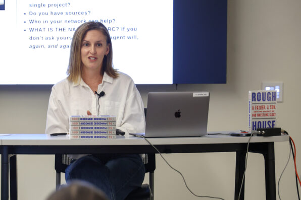 A woman speaks in front of a digital screen. There is a stack of books titled "Rough House" in front of her.