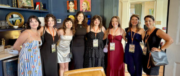 A group of eight women stand together in dresses wearing lanyards, smiling widely in a blue, wooden room.