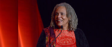A picture of Charlayne Hunter Gault speaking at a formal even with red and black piping in the background.