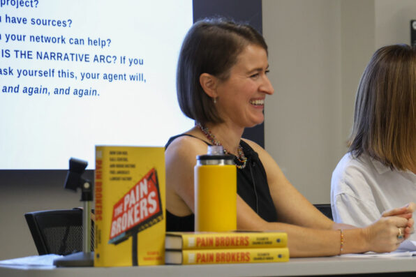 Elizabeth Chamblee Burch sits at a table and smiles. Her book, "Pain Brokers" is propped up on the table in front of her.