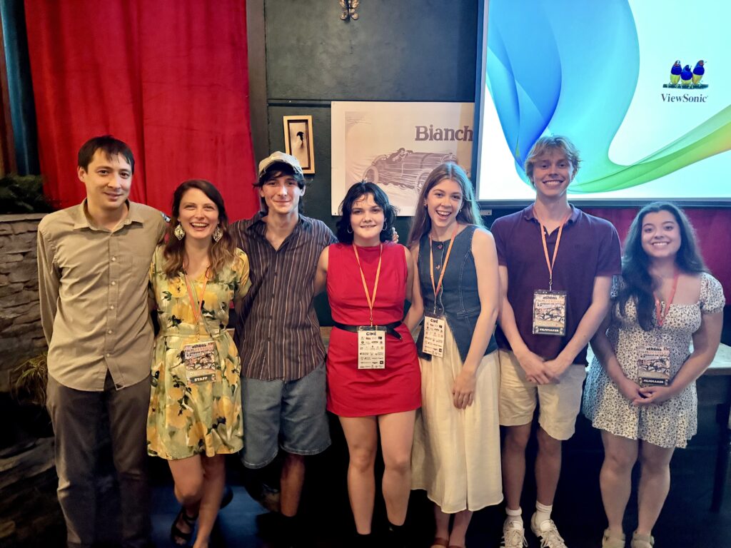 A group of seven people, three boys and four girls, stand in a room with a screen projector and red drape while they wear various levels of attire. 