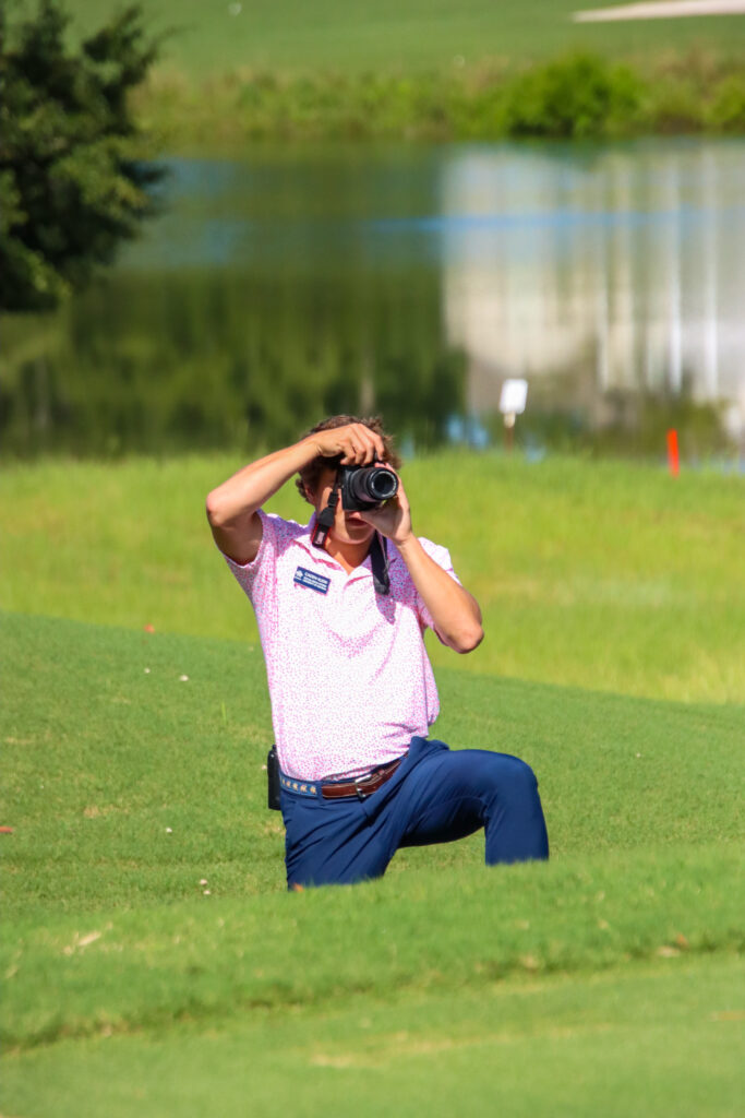Caden Klein kneels on a golf course with a camera while covering an event.