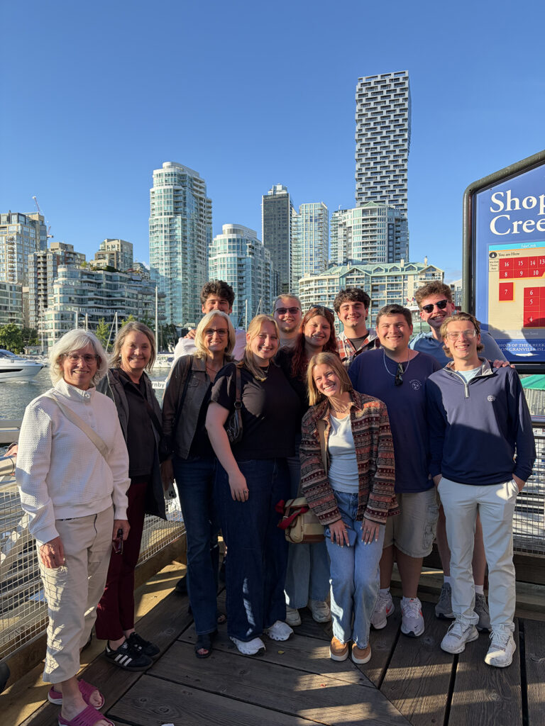 A group of women and students pose for a picture on a pier. Several modern skyscrapers are in the background.