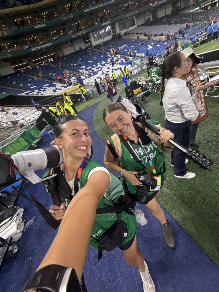 Two female photographers in photo bibs and carrying camera gear pose for a selfie in the sidelines of a large stadium.