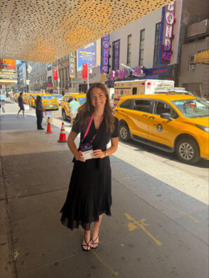 Elizabeth Nark smiles holding a badge in front of taxis on a New York City street.