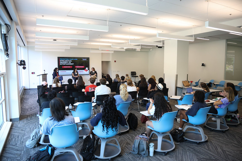 A room full of students sit in chairs, listening to three women speak in the front of the room.
