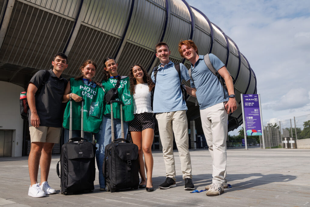 A group of six students, two with photographer's bibs and rolling bags in front, pose for a picture outside of a large stadium.