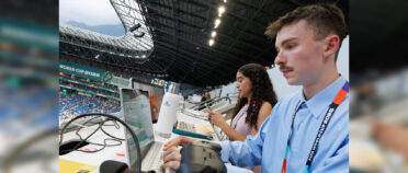 A close-up of a female and male students sitting at their computers in a large open-air stadium. There is a sign in the background that reads World Cup 2026.