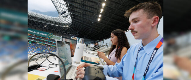 A close-up of a female and male students sitting at their computers in a large open-air stadium. There is a sign in the background that reads World Cup 2026.