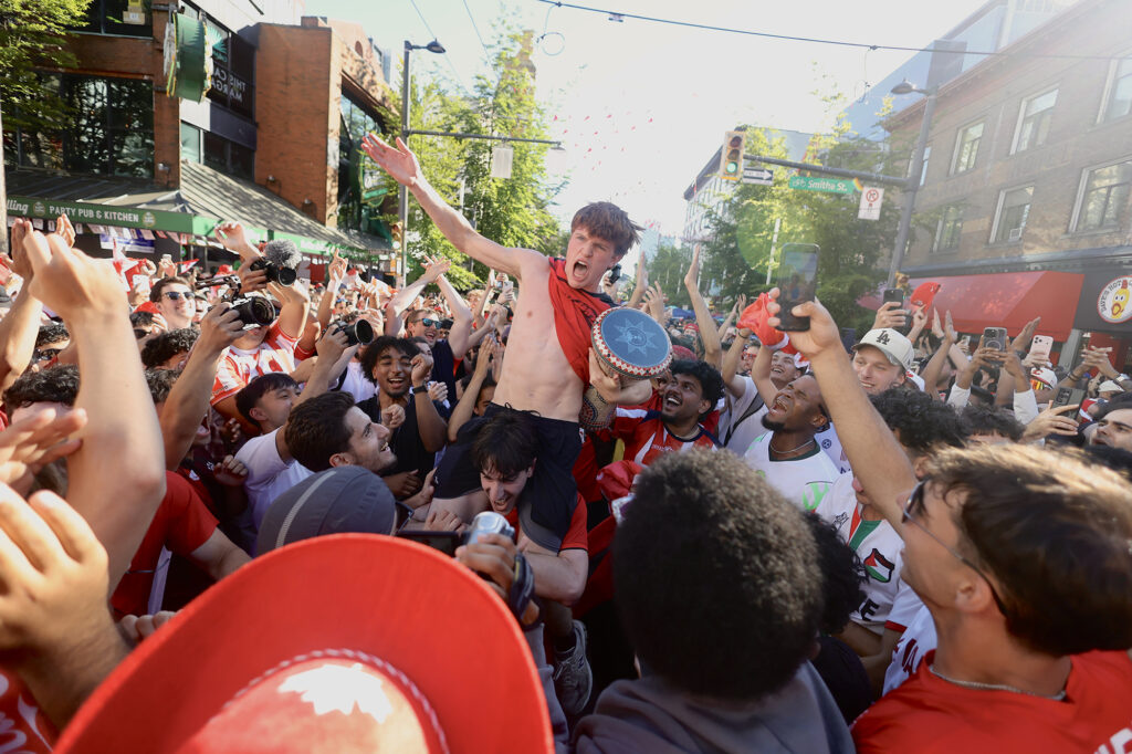 A young man with his shirt hanging around his neck is hoisted on the shoulders of other fans parading outside in a street.