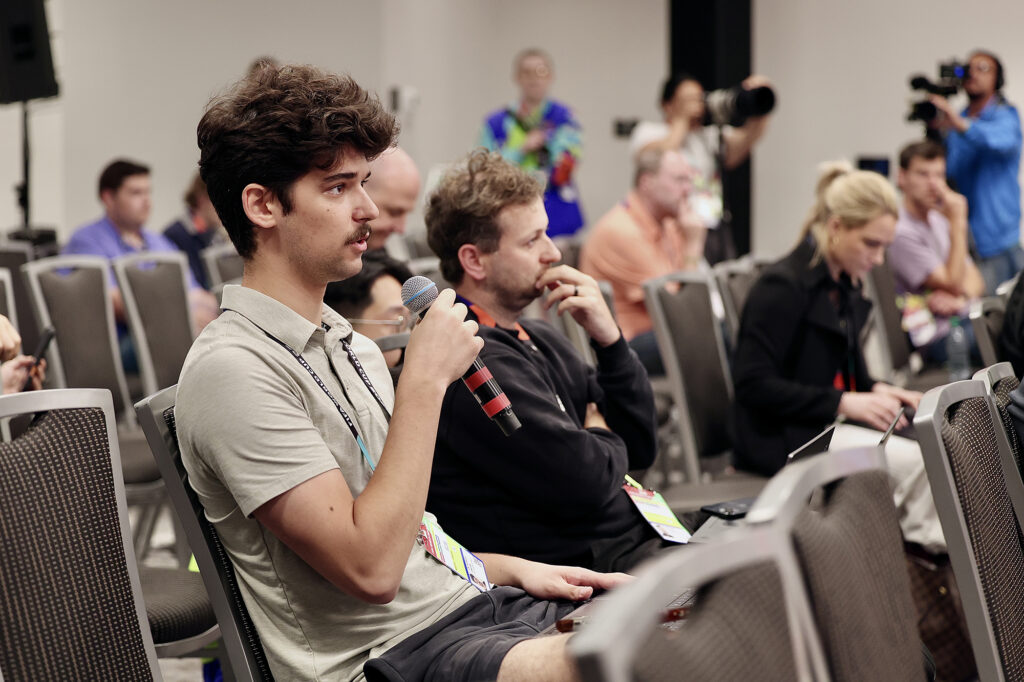 Close up profile of a student holding a microphone asking a question in a group of people sitting down. Cameras are out of focus in the background.