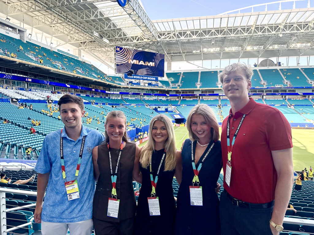 Five students wearing press credentials pose for a picture with a large stadium in the background. The digital screen behind reads Miami.