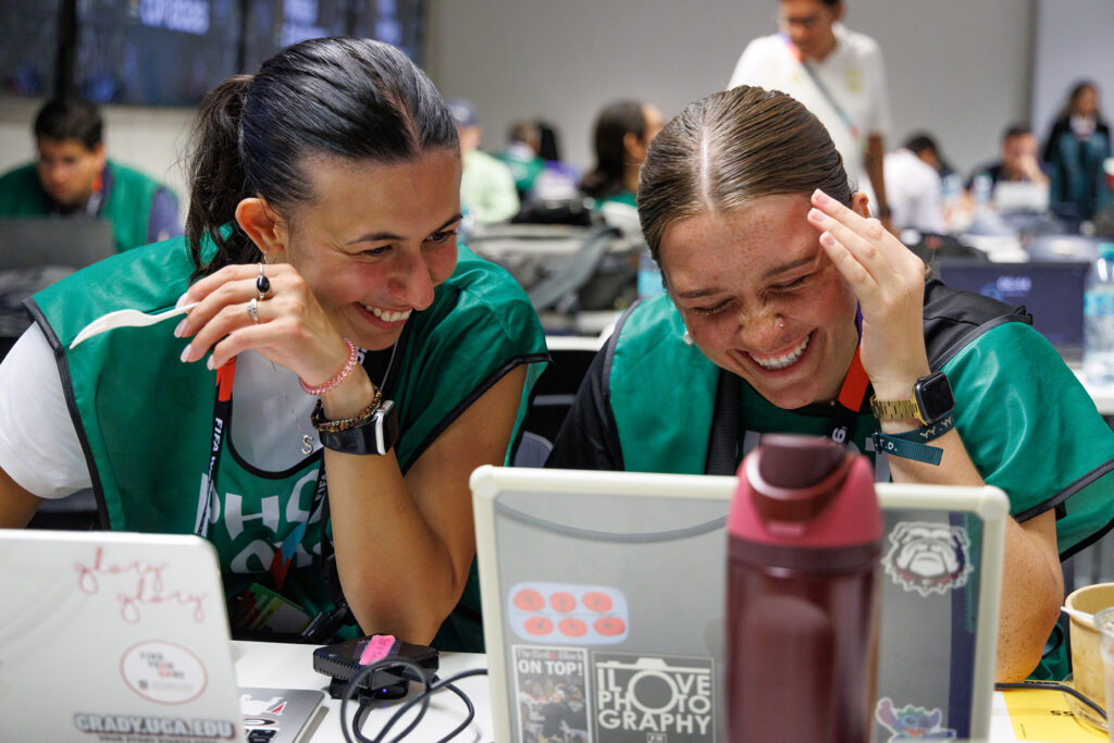 Two females sit at computers and laugh. They are wearing green photographer's bibs.