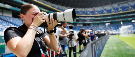 A female photographer has a long-lens camera and takes a pictures with a large stadium and field in the background.