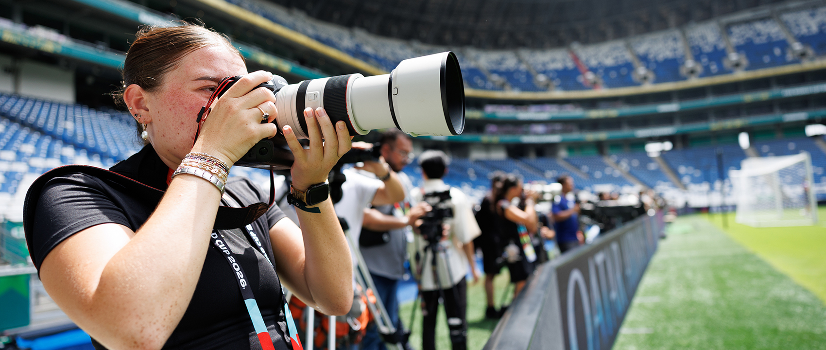 A female photographer has a long-lens camera and takes a pictures with a large stadium and field in the background.