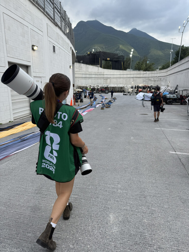 The back view of a woman walking in the outside part of a stadium with a mountain in the background. She is carrying a long lens camera and a photographer's photo bib.