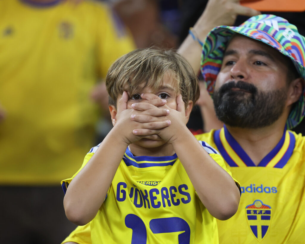 A young boy wearing a yellow soccer jersey covers his mouth in disbelief while watching a match. A man with a beard and hat sits behind him.