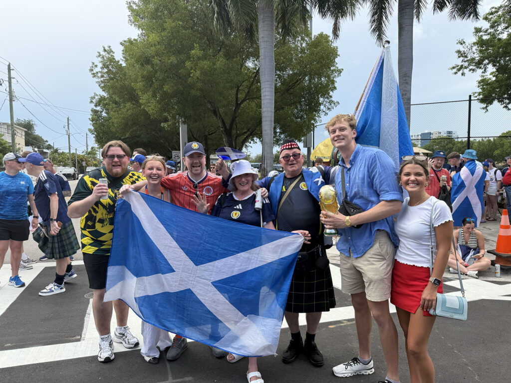 Scottish fan in kids and hats pose for a picture outside with students. They are holding large blue flag with a white X.