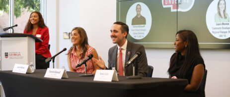 A man and two women sit at a table with microphones addressing an audience. A student in a red blazer looks on.