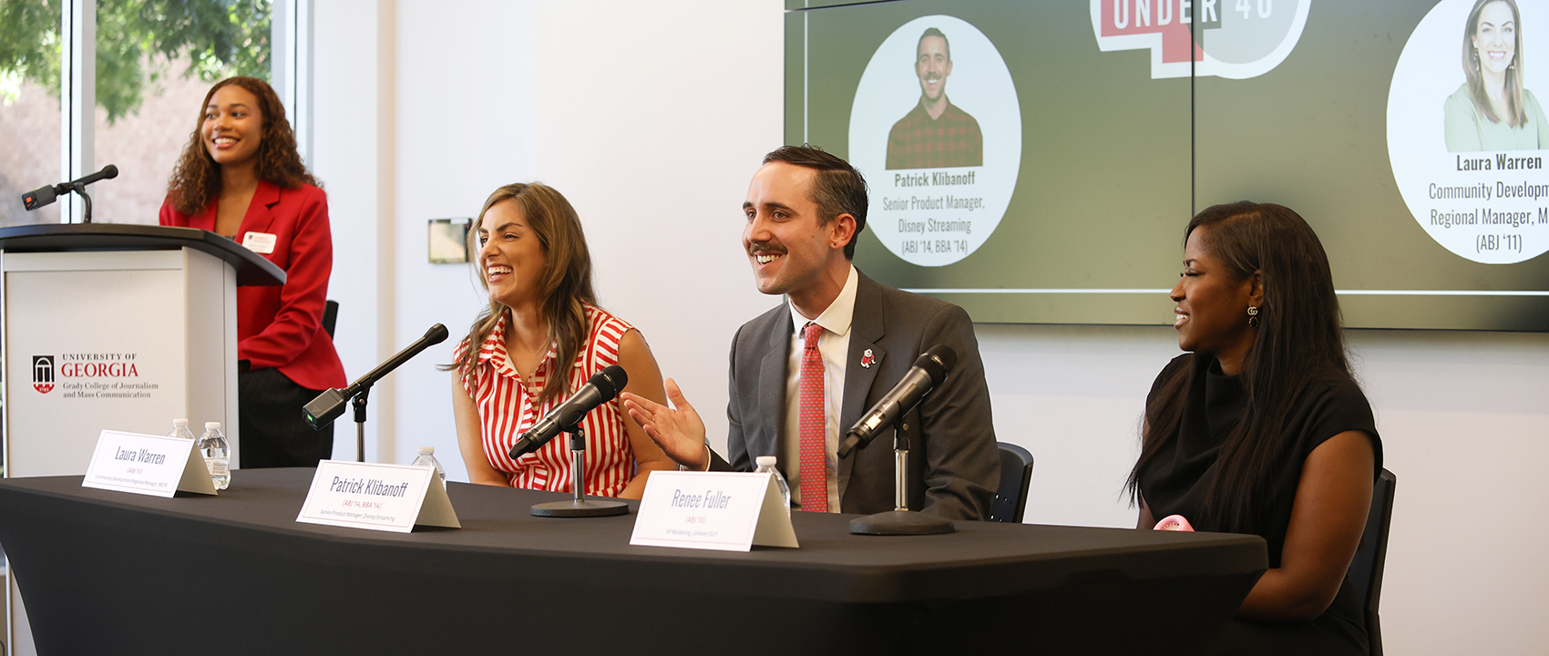 A man and two women sit at a table with microphones addressing an audience. A student in a red blazer looks on.