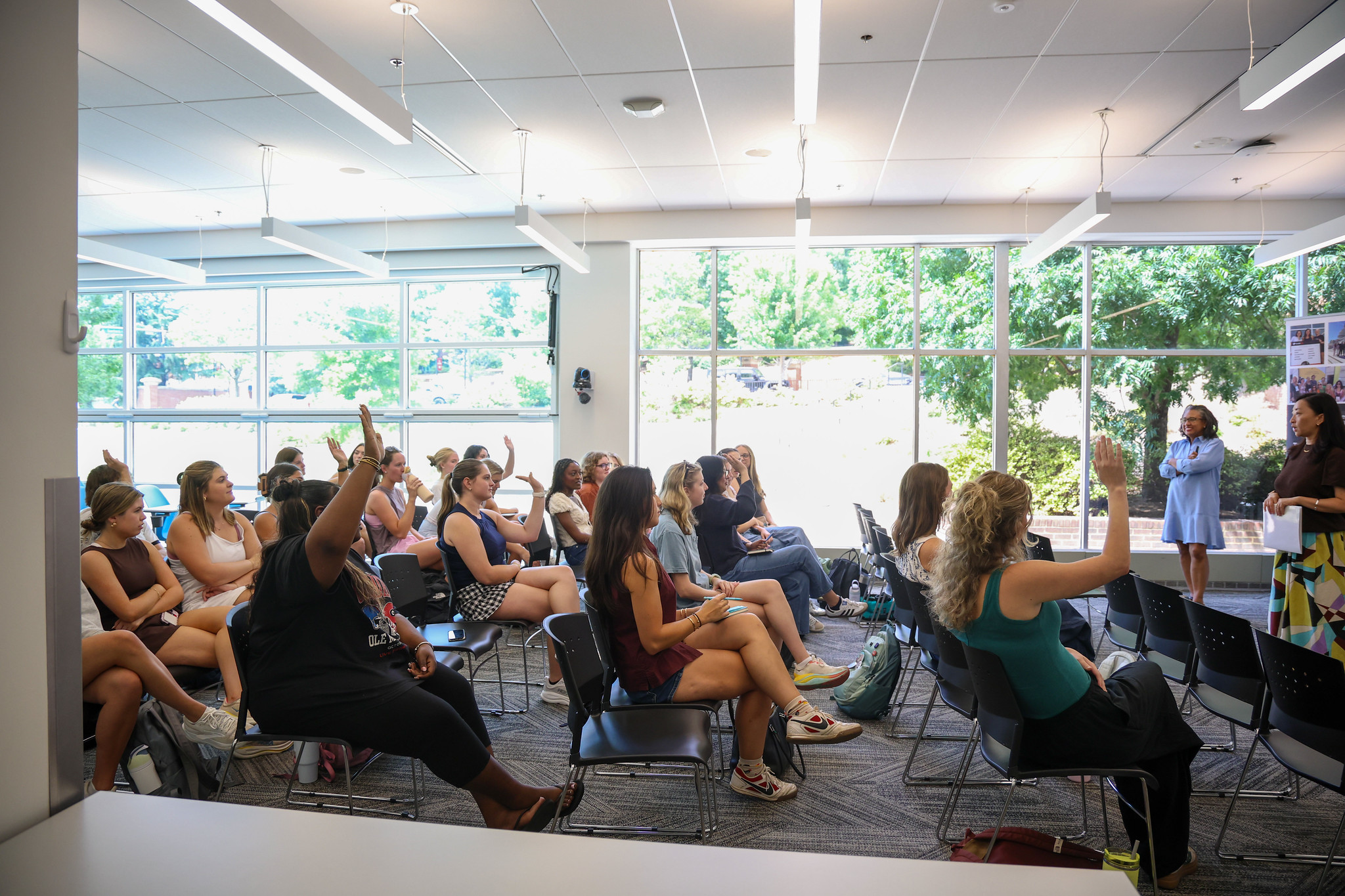 A profile view of a group of students, some raising their hands, in a lecture with two woman standing in the front of the room.