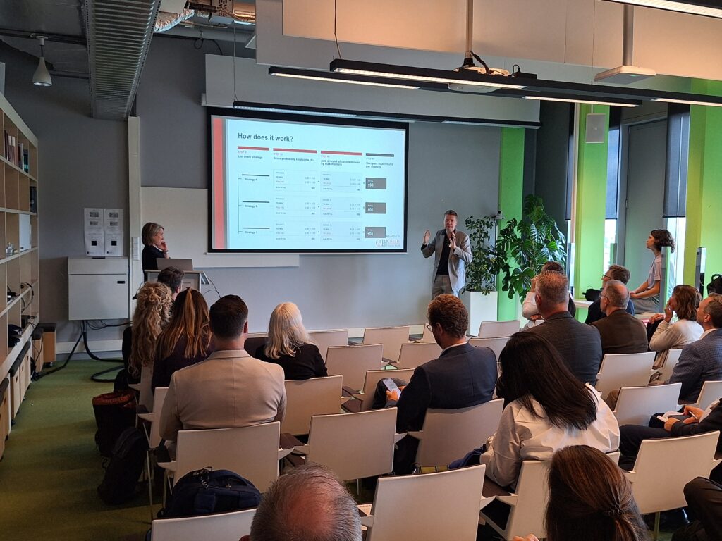 A man presents to a group of people. The digital screen behind him has the title how does it work?