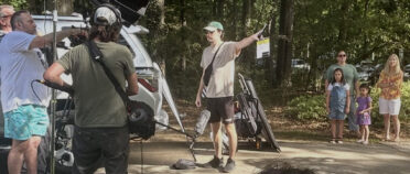 A man holds a film slate as a camera crew gets ready to film a family, seen in the background.