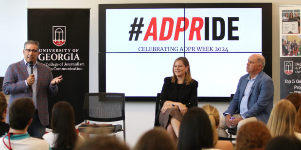 A man holding a microphone and a man and woman sitting talk with a large group of students. A digital projection in the background says #ADPRIDE.