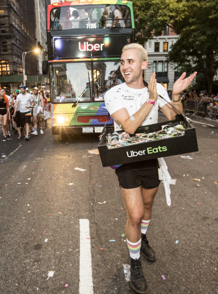 Patrick Klibanoff marches in New York City's Pride Parade while handing out goodies from UberEats. Behind him is an Uber-branded bus painted pride flag colors.