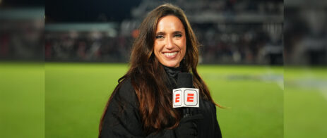 A woman dressed in a black coat smiles while holding a microphone with an E on it. She is standing on an athletic field.