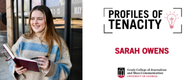 Sarah Owens smiles holding a red notebook and pen outside of a black glass building.