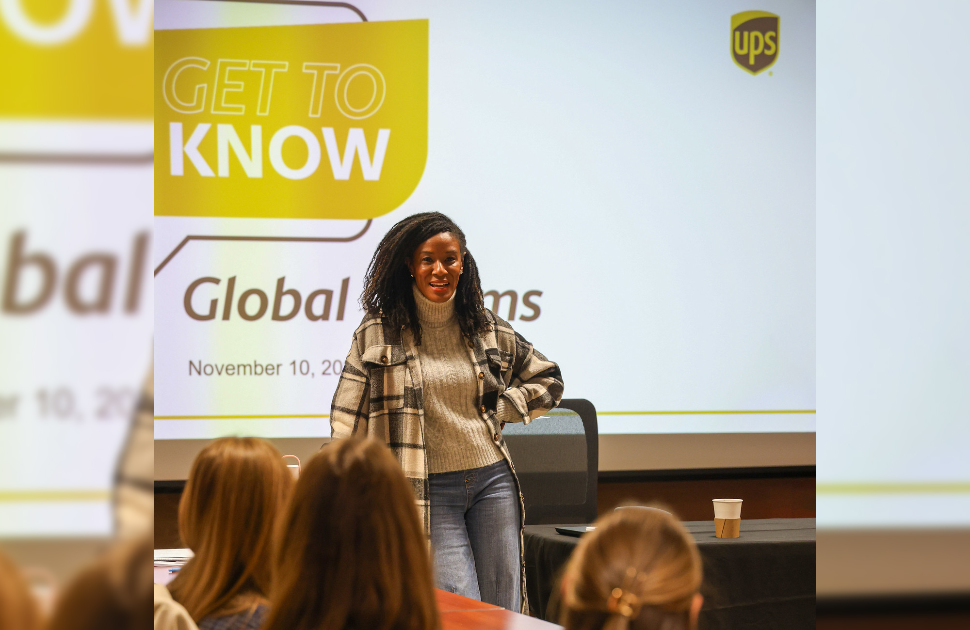 A woman stands and talks with some students. In the background is a digital screen that says UPS and Get to Know.