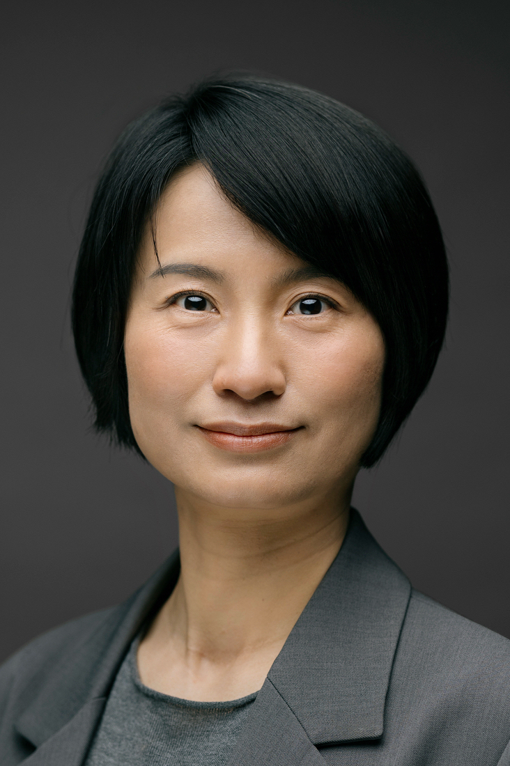 A person with short black hair, wearing a gray blazer and shirt, looks confidently at the camera against a plain dark background.