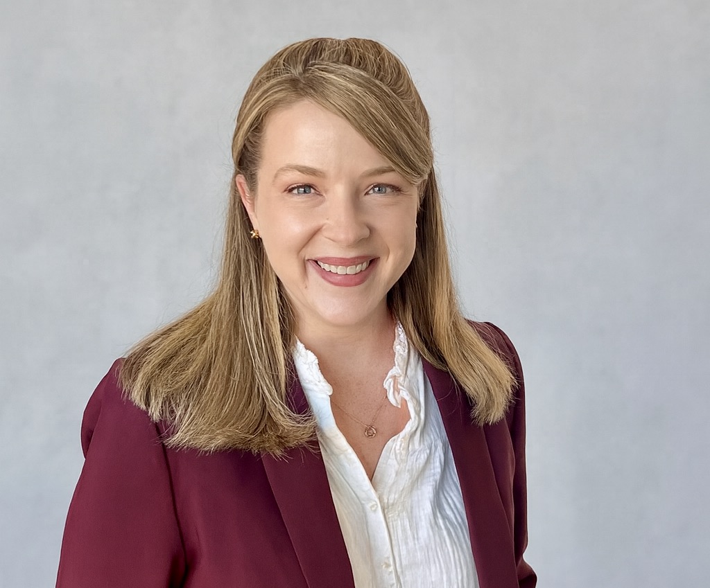 A smiling woman with blonde hair, wearing a white button-down shirt under a dark red blazer.