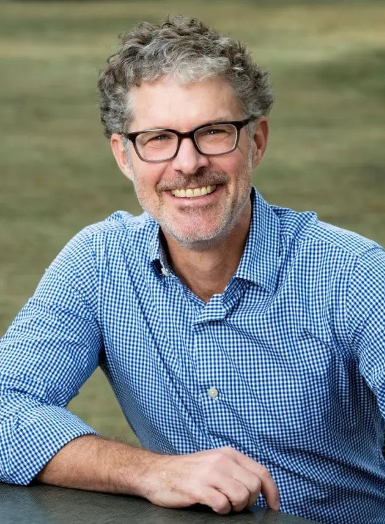 A middle-aged man with curly salt-and-pepper hair, glasses, and a beard smiling while wearing a blue gingham button-down shirt.