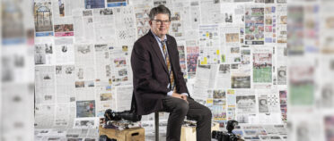 Mark Johnson sits on a stool in front of a backdrop with newspapers. He has a few cameras on wooden boxes at his feet.