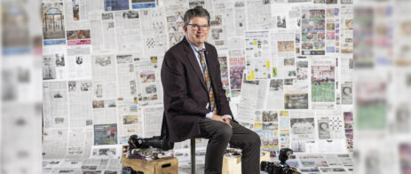 Mark Johnson sits on a stool in front of a backdrop with newspapers. He has a few cameras on wooden boxes at his feet.