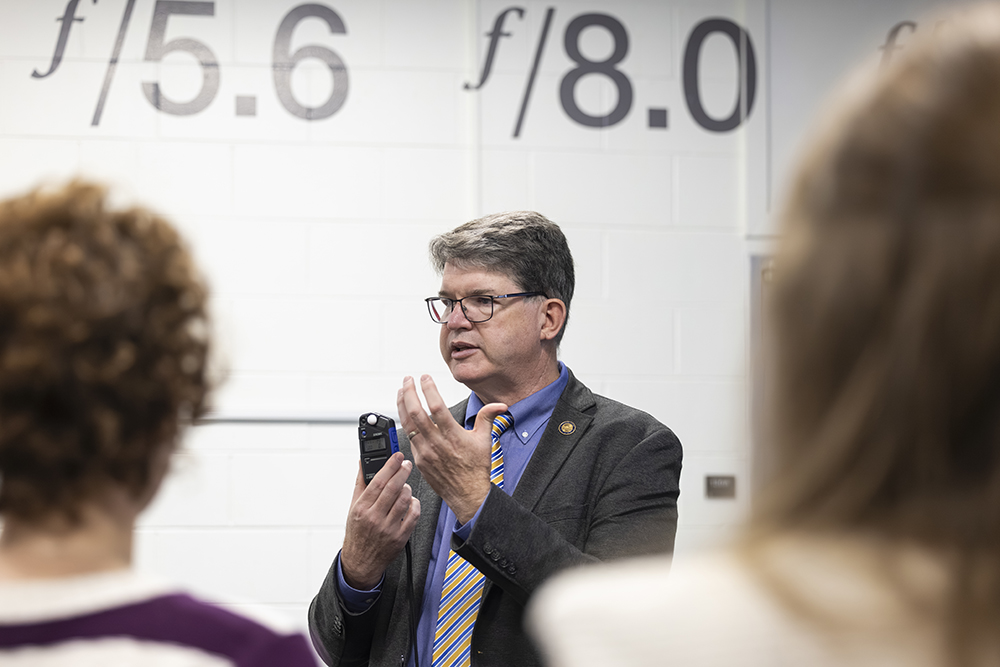 Mark Johnson holds a light meter in his hand and gestures with the other hand. 